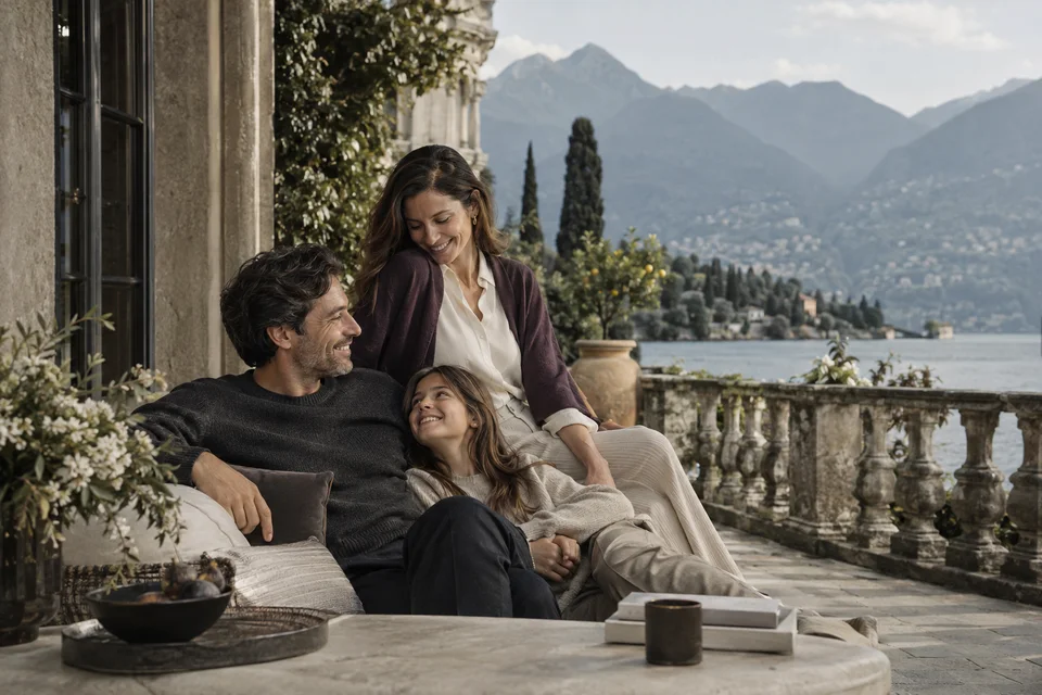 Família em uma varanda de pedra com vista para um lago e montanhas, em uma cena serena de viagem.